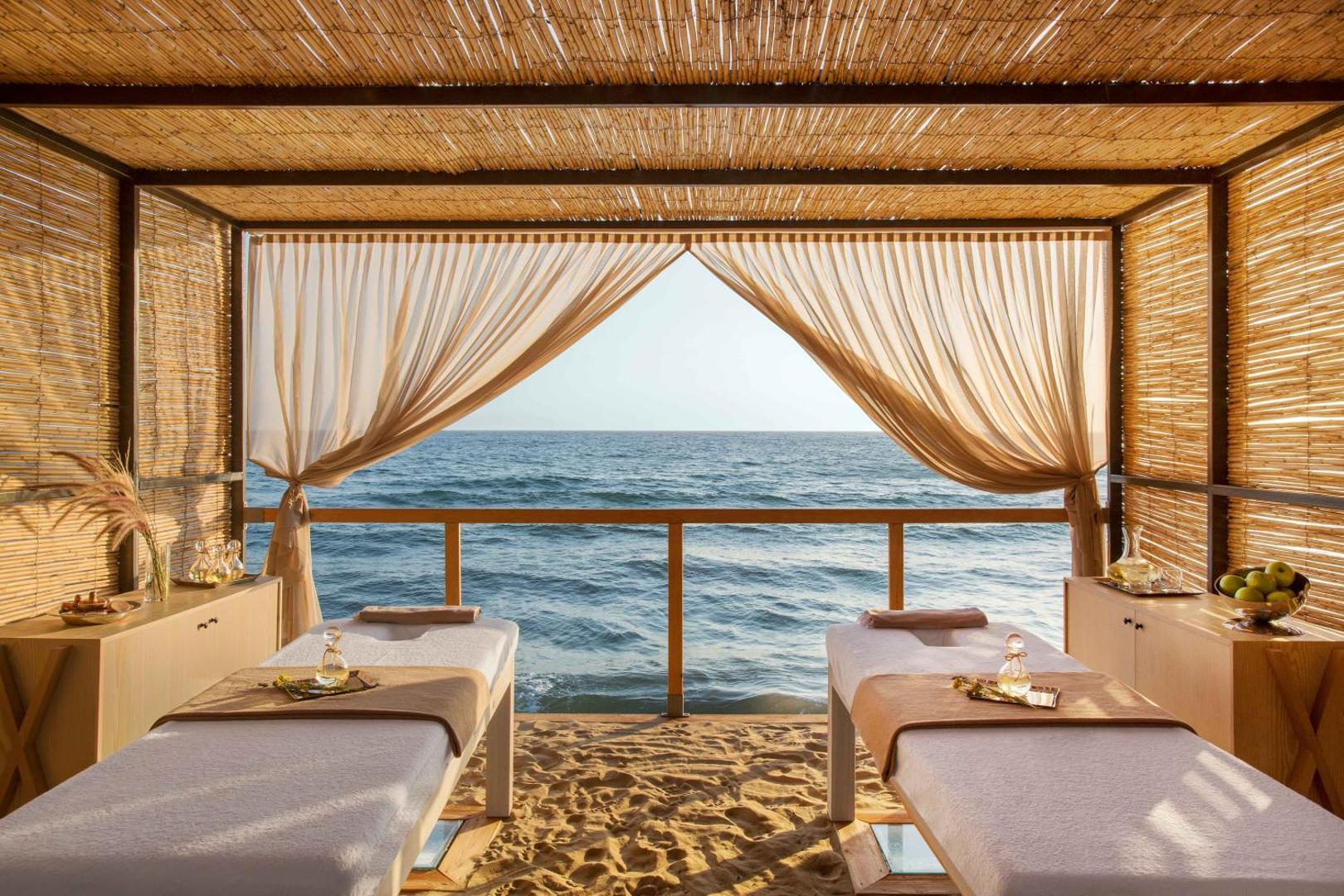 Club Marvy spa procedūru zona pludmalē ar skatu uz jūru — divi masāžas galdi zem bungalo tipa nojumes smilšainā krastā