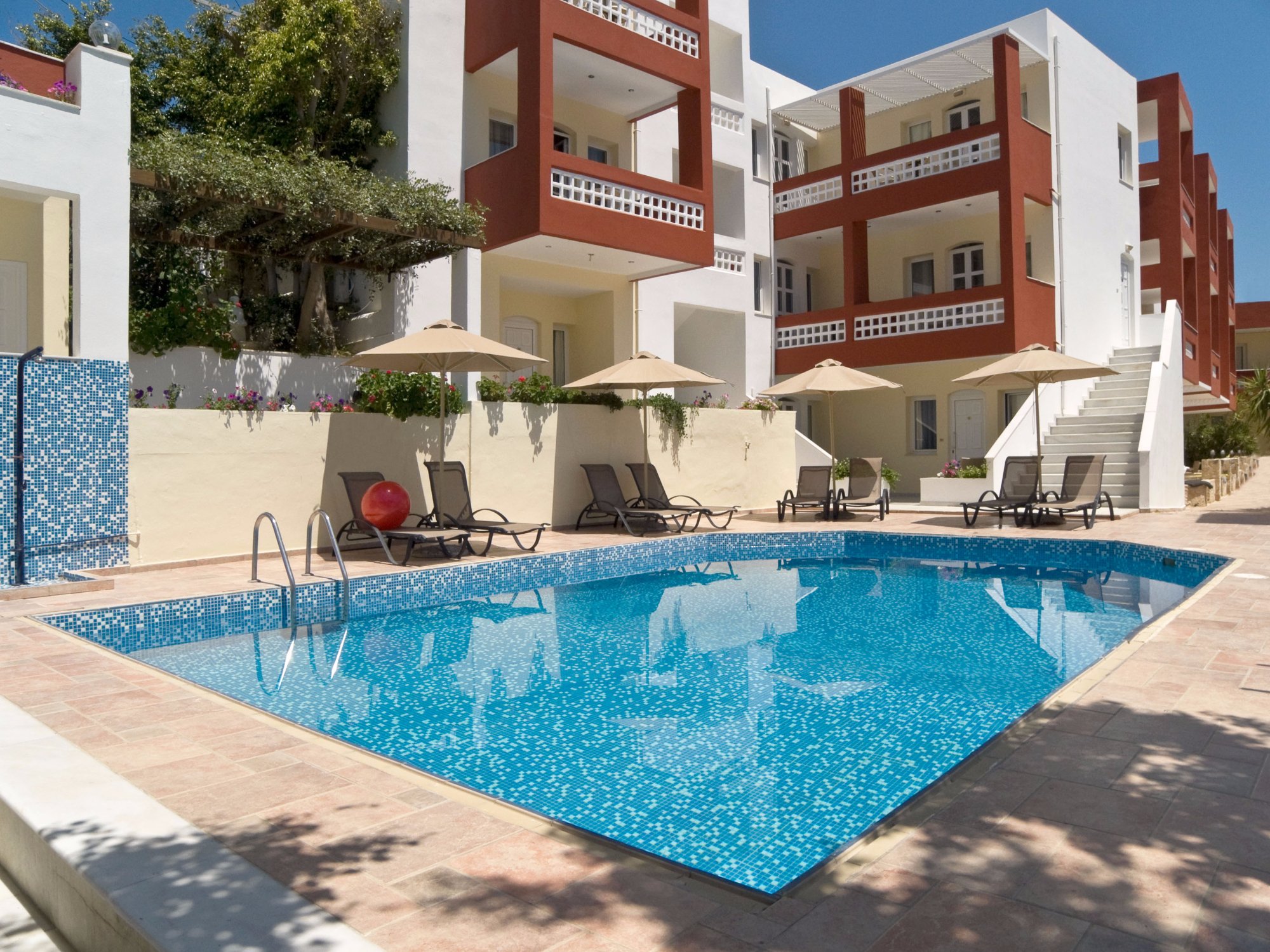 Troulis Apart-Hotel iekšpagalma baseins ar zilu mozaīku — apkārt sauļošanās krēsli, saulessargi un viesnīcas korpusi ar balkoniem Bali, Krētā.