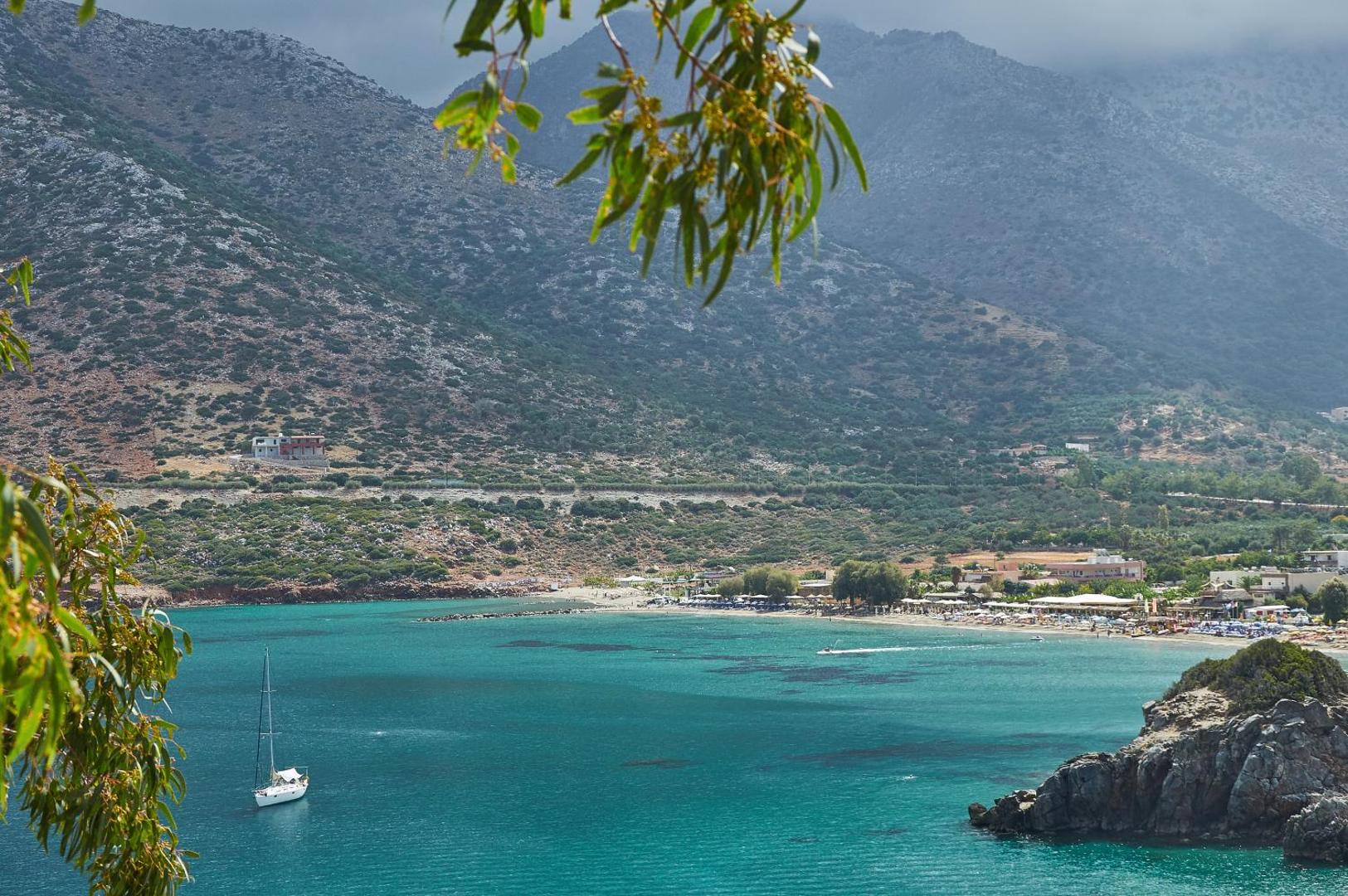 Troulis Apart-Hotel skats uz tirkīzzilo Bali līča pludmali Krētā — kalnu ainava, jahta un piekrastes atpūtas zona caur koku lapotni