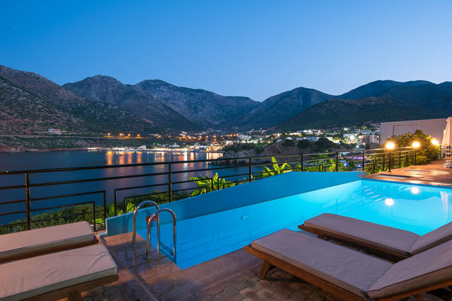 Troulis Apart-Hotel baseins ar bezgalības malu vakarā — skats uz Bali ciemata līci un kalniem Krētā, izgaismoti sauļošanās krēsli.