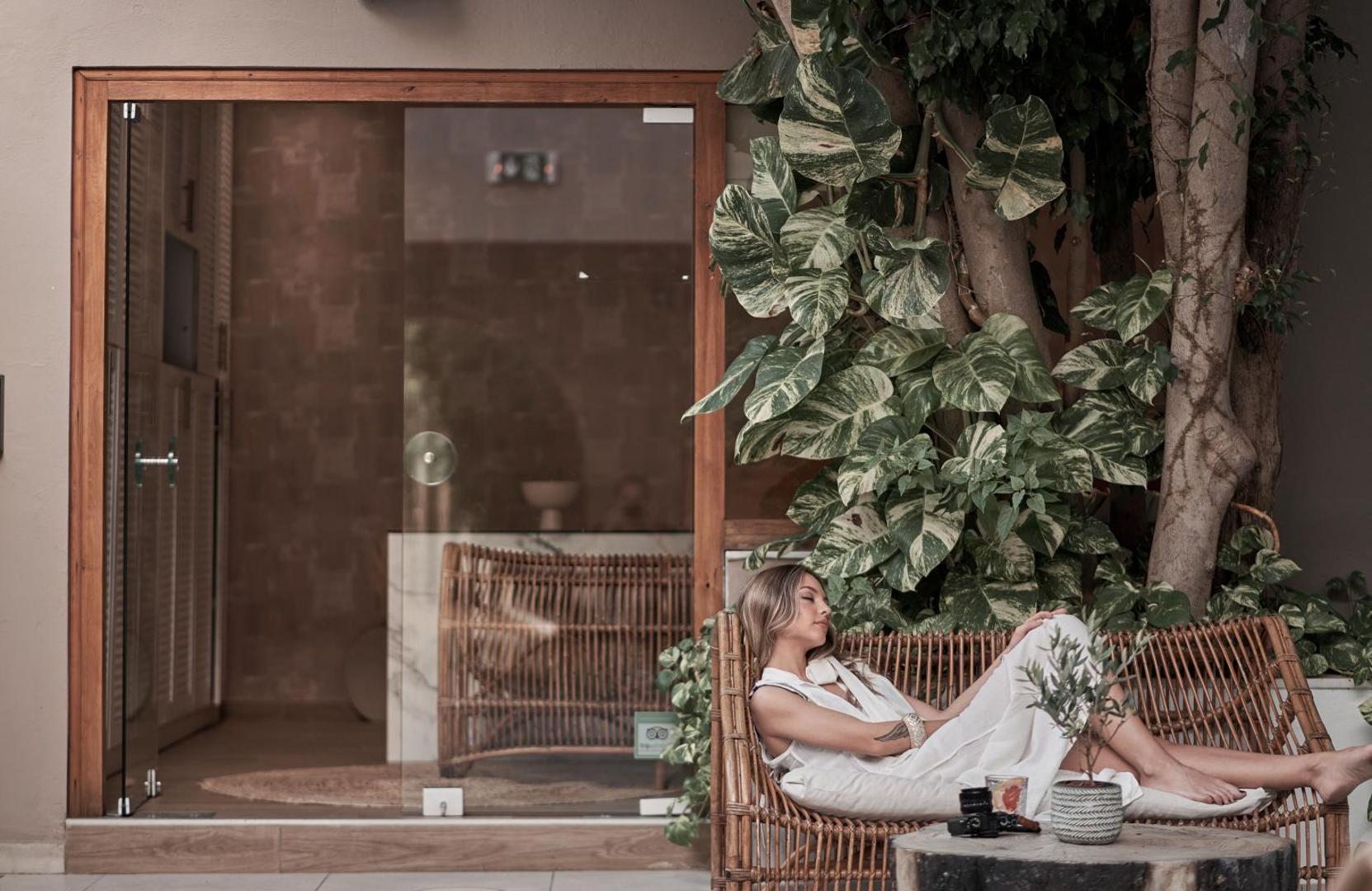 Viesis atpūšas Troulis Apart-Hotel āra terasē uz pīta dīvāna pie zaļojoša koka blakus viesnīcas ieejai Bali kūrortā