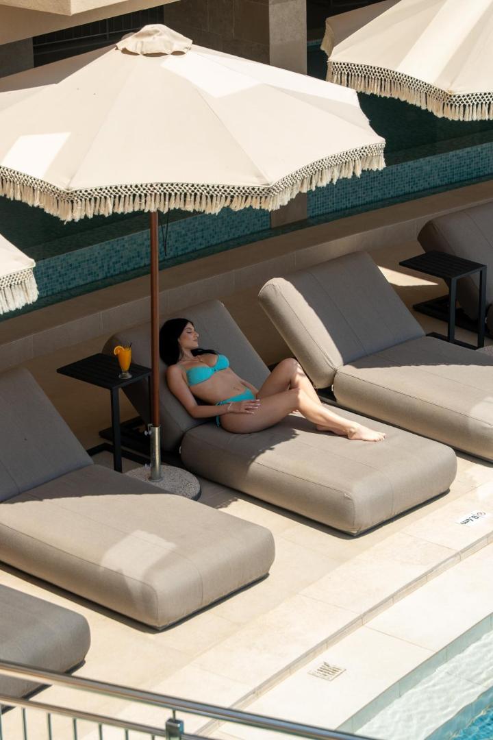 Viesis atpūšas uz mīksta sauļošanās krēsla zem saulessarga pie Ventale Island Breeze Resort baseina Krētā — vasaras atpūta ar kokteili.