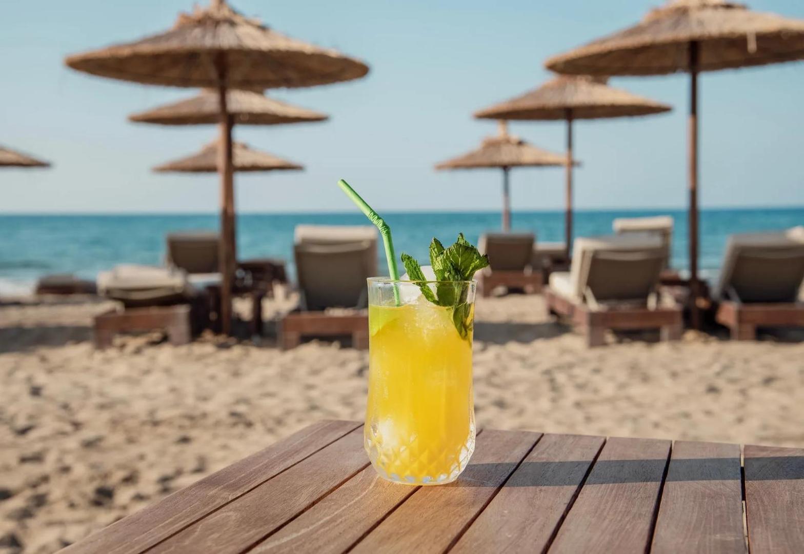 Ventale Island Breeze Resort privātā pludmale ar saulessargiem un zviļņiem, priekšplānā atspirdzinošs dzēriens uz koka galdiņa pie jūras.