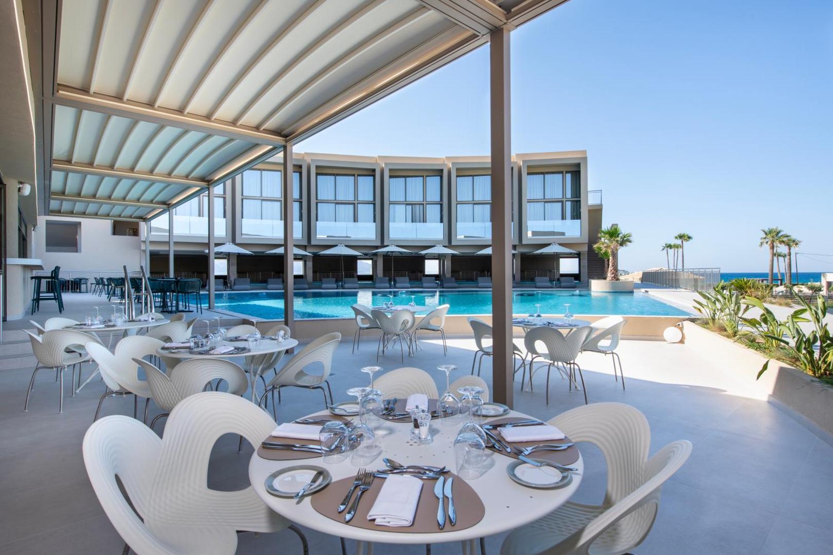 Ventale Island Breeze Resort restorāna terase ar dizaina krēsliem pie baseina un skatu uz viesnīcas moderno arhitektūru