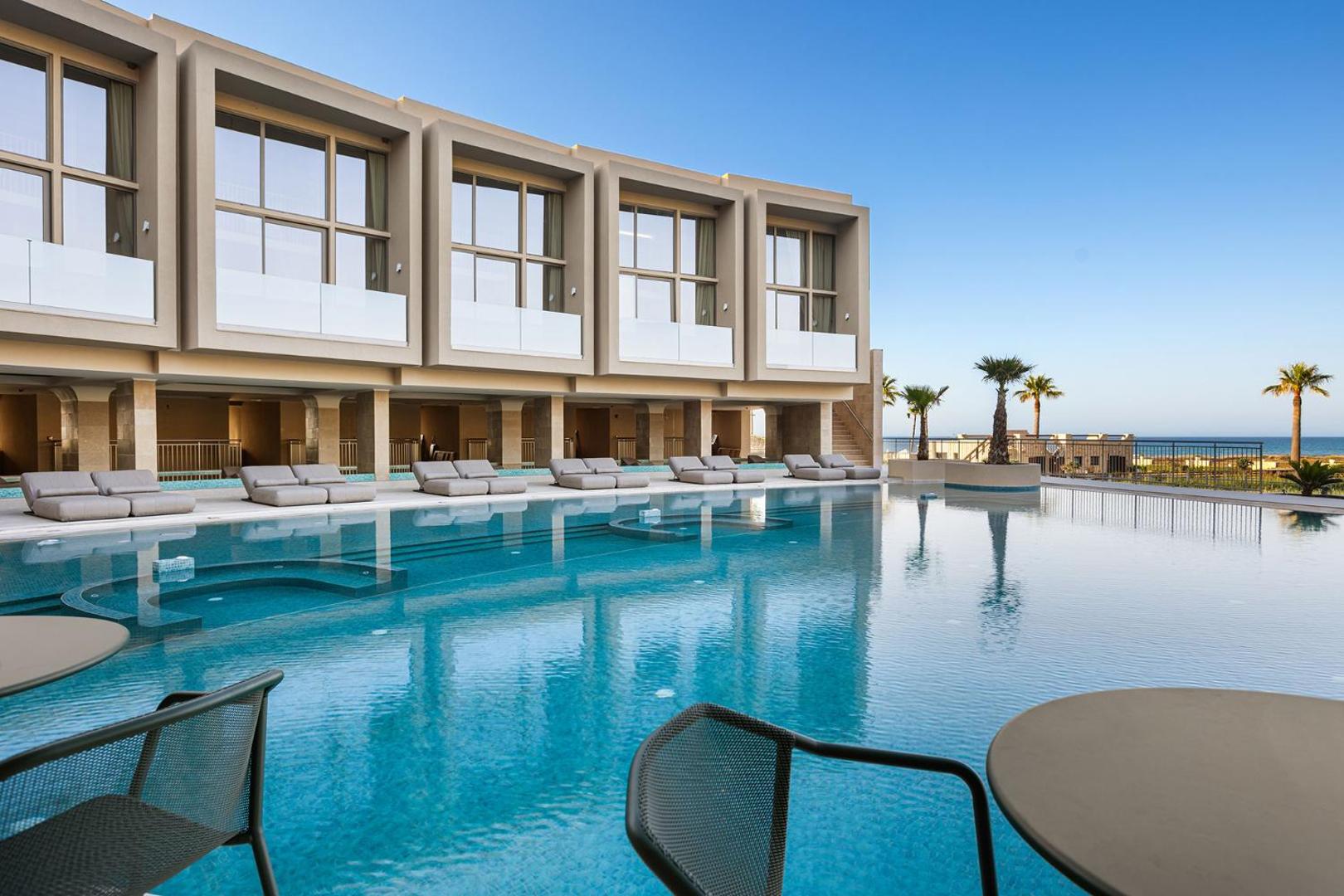 Ventale Island Breeze Resort galvenais āra baseins ar sauļošanās krēsliem un skatu uz jūru piekrastē, Krētā — moderna arhitektūra un palmas.