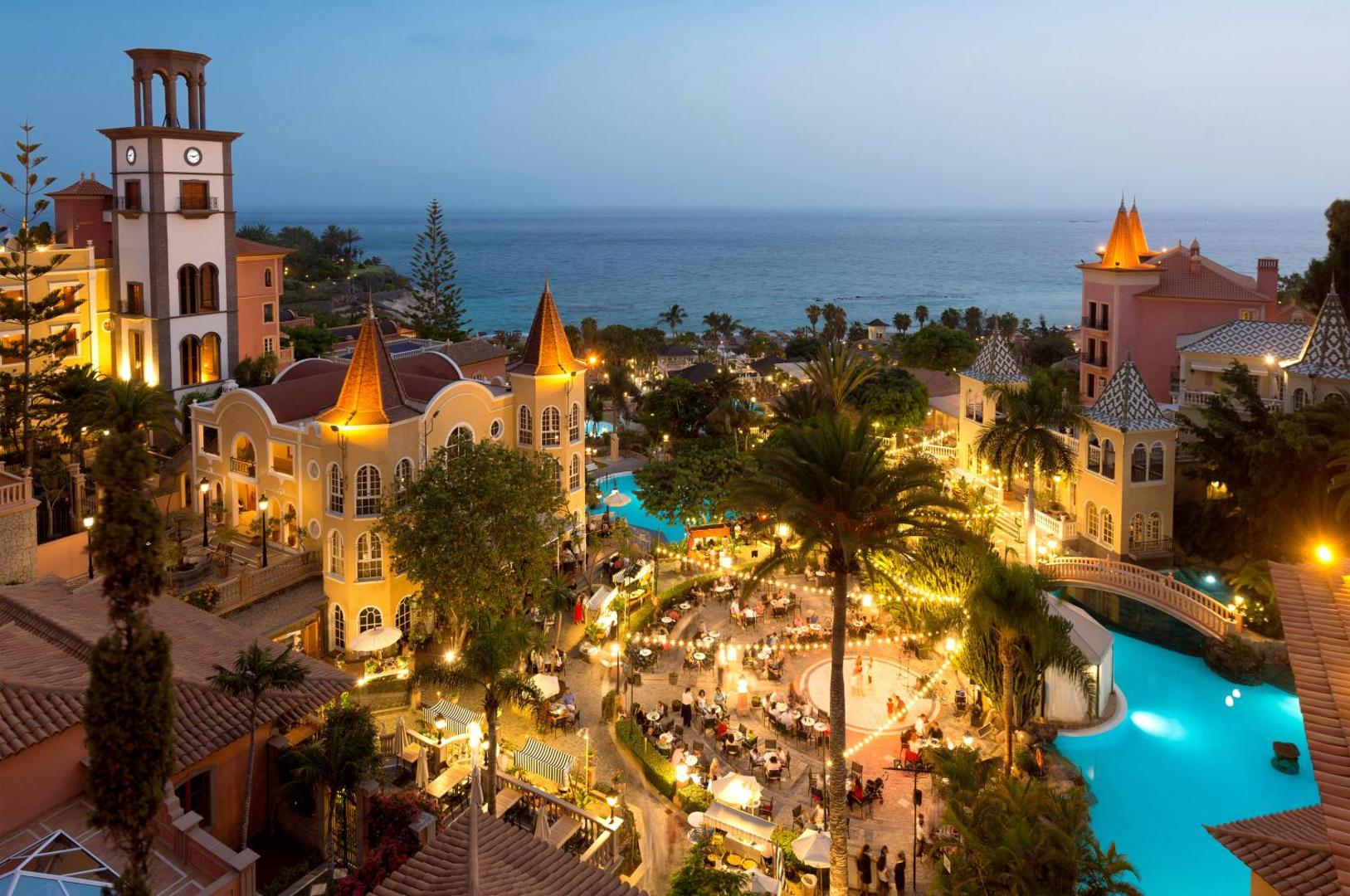 Bahia del Duque kūrorta vakara panorāma Tenerifē ar izgaismotiem baseiniem, restorāna terasi un skatu uz okeānu saulrietā.