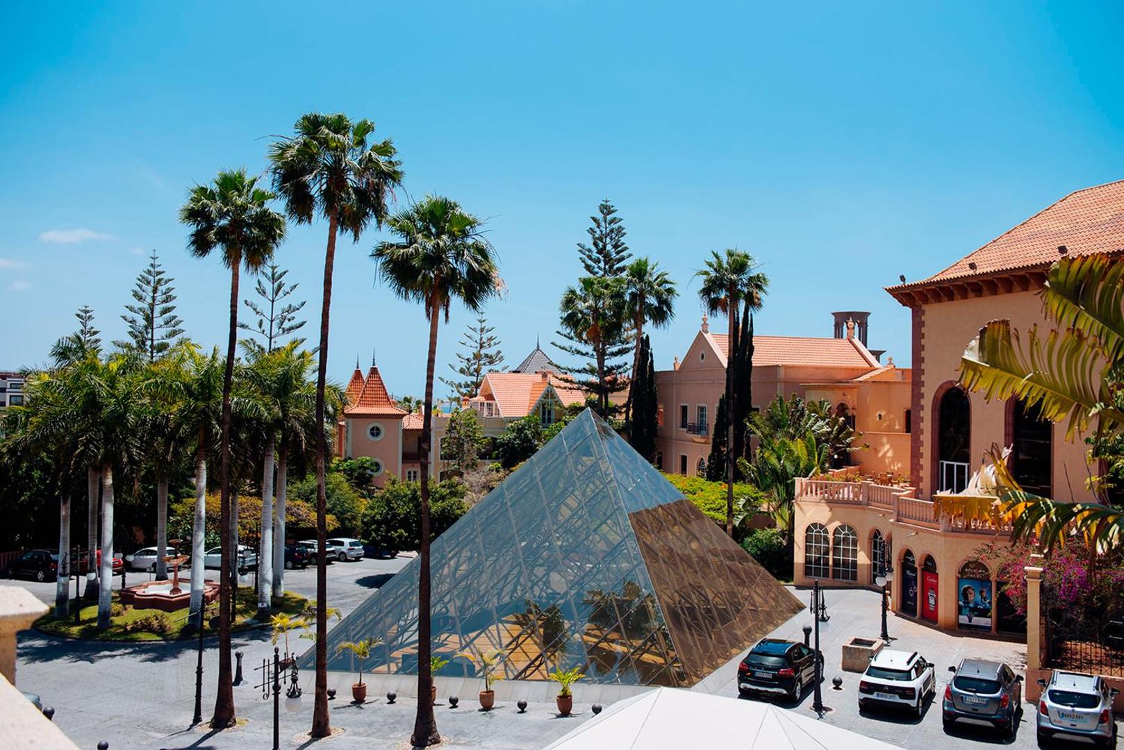 Bahia del Duque ieejas zona ar modernu stikla piramīdu, palmām un klasiskā stila viesnīcas korpusiem saulainā dienā.