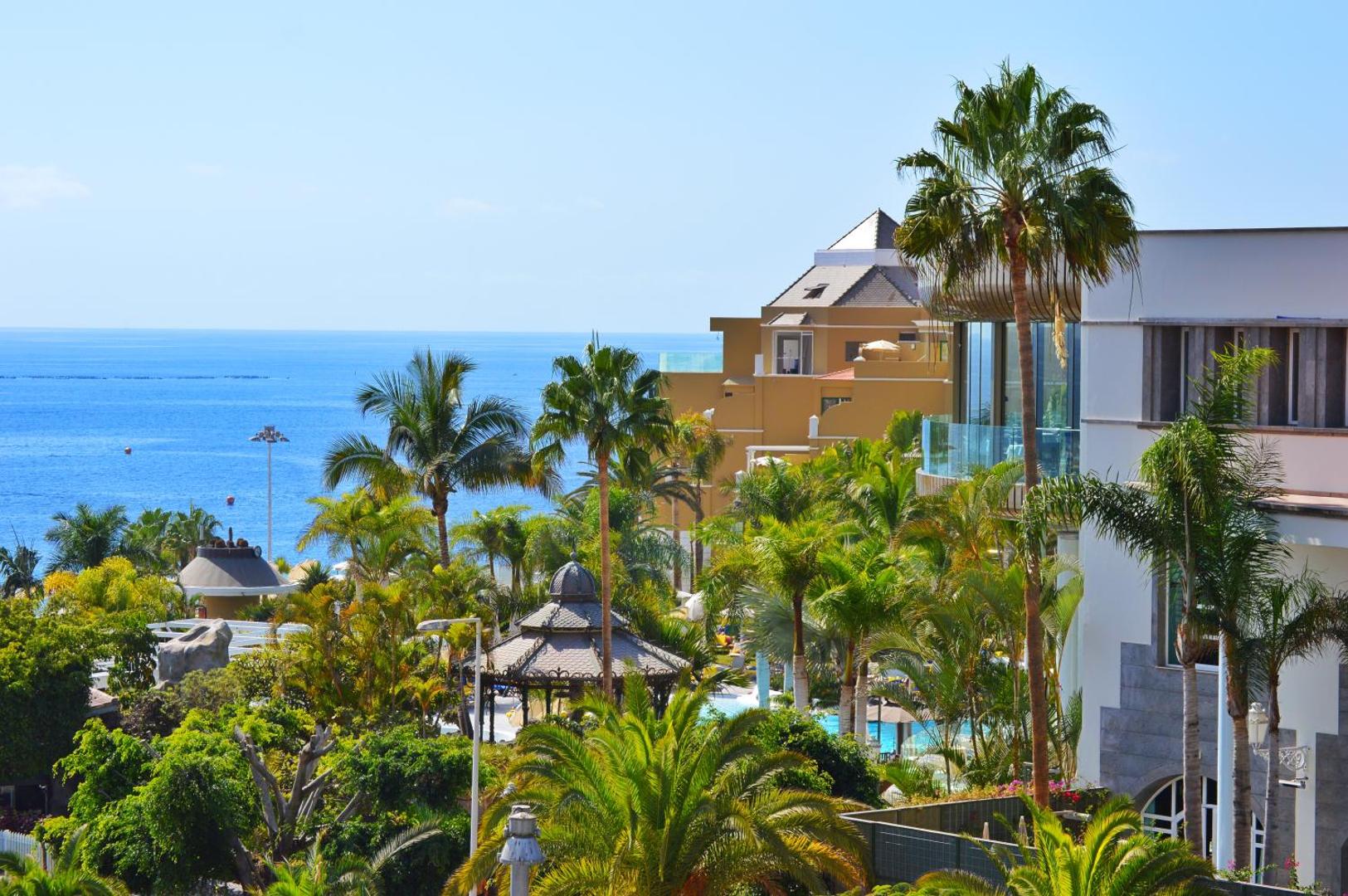 Skats no Jardines de Nivaria viesnīcas terases uz zilo okeānu caur lekniem palmu dārziem un kūrorta arhitektūru Tenerifes dienvidos.