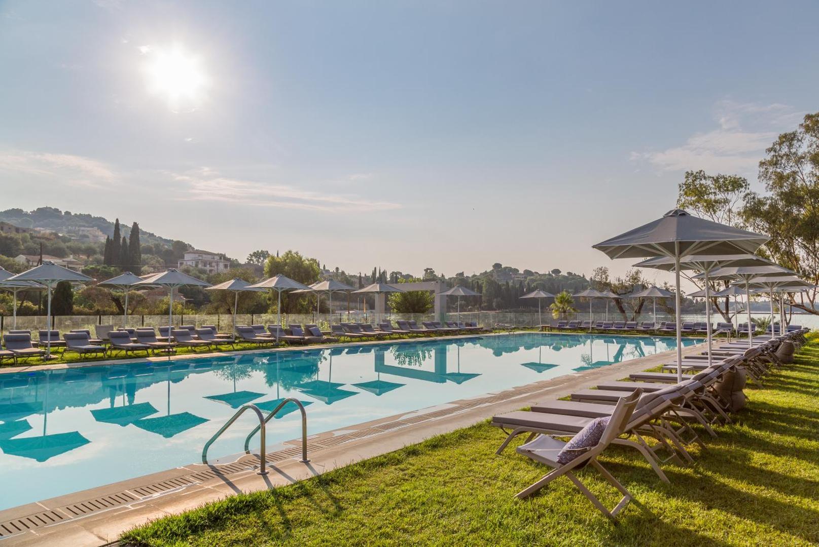 Rodostamo Hotel & Spa plašais āra baseins saulrietā ar zvilnētājiem, saulessargiem un skatu uz Korfu kalniem un zaļumiem