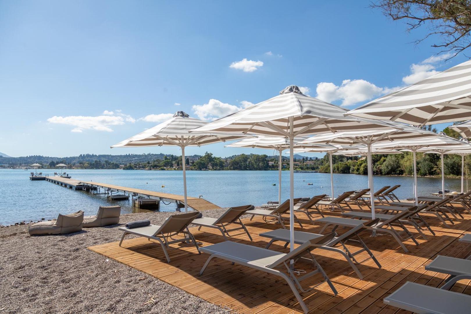 Rodostamo Hotel & Spa privātā pludmales zona ar koka klāju, saulessargiem, zviļņiem un garu laipu jūrā Korfu salā