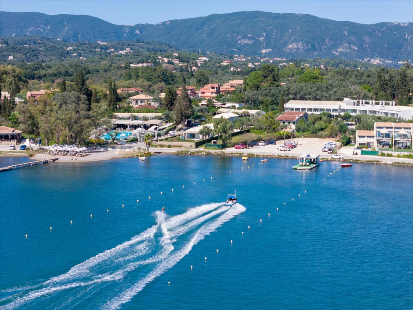 Aerofoto skats uz Rodostamo Hotel & Spa piekrasti Korfu — zils līcis ar motorlaivu un viesnīcas kompleksu fonā