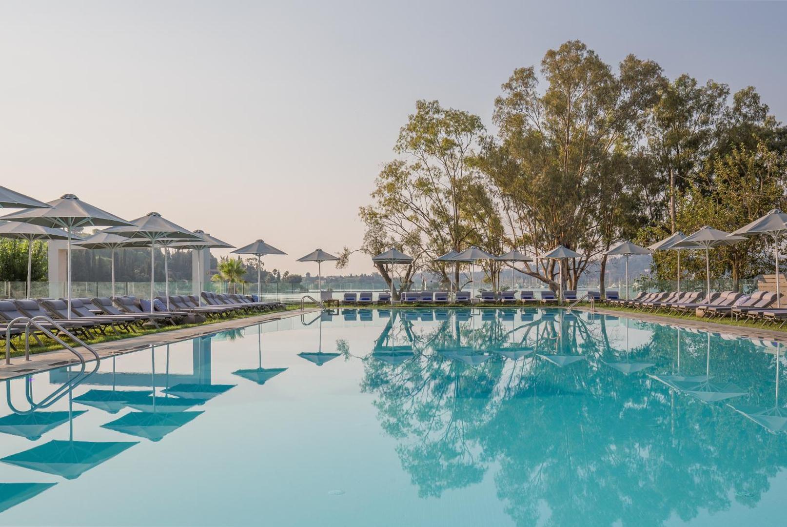 Rodostamo Hotel & Spa galvenais baseins ar spoguļattēlu ūdenī, apkārt rindojas pelēki saulessargi un koki piekrastē