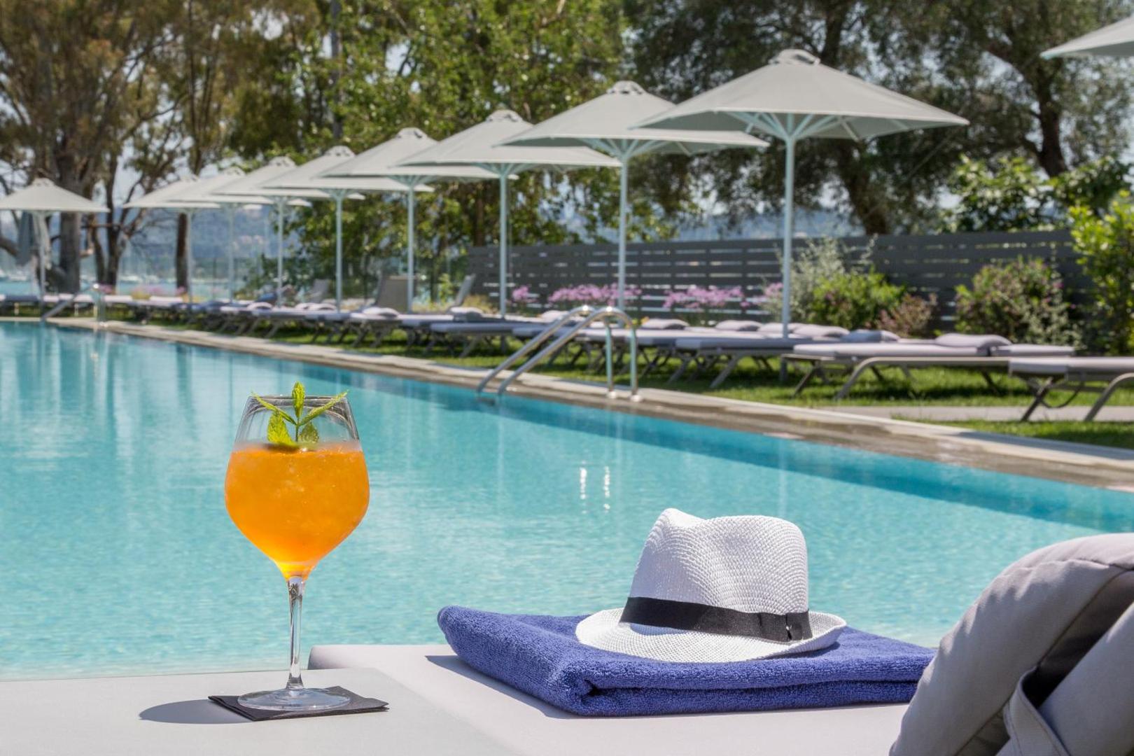 Atsvaidzinošs kokteilis, cepure un dvielis uz zviļņa pie Rodostamo Hotel & Spa baseina, fonā saulessargi un koki