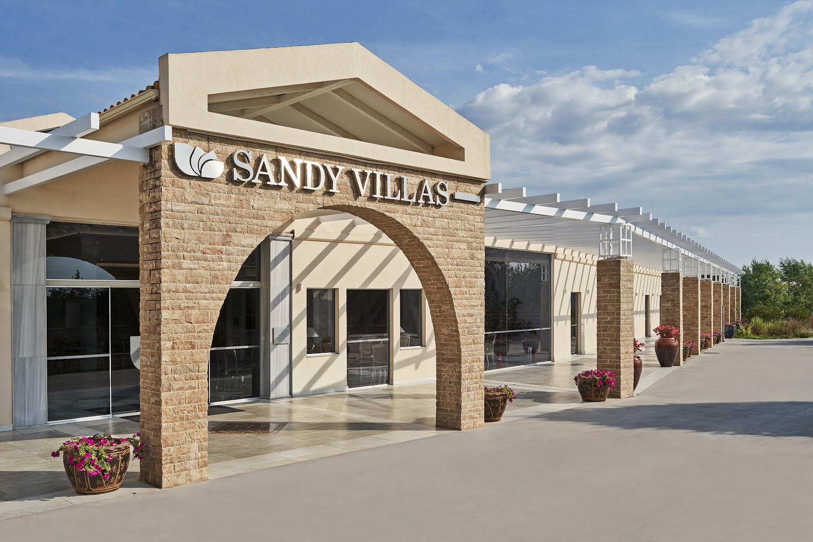 Sandy Villas Resort galvenā ieeja ar akmens arkām un ziedu podiem saulainā dienā — moderna arhitektūra Grieķijas piekrastē
