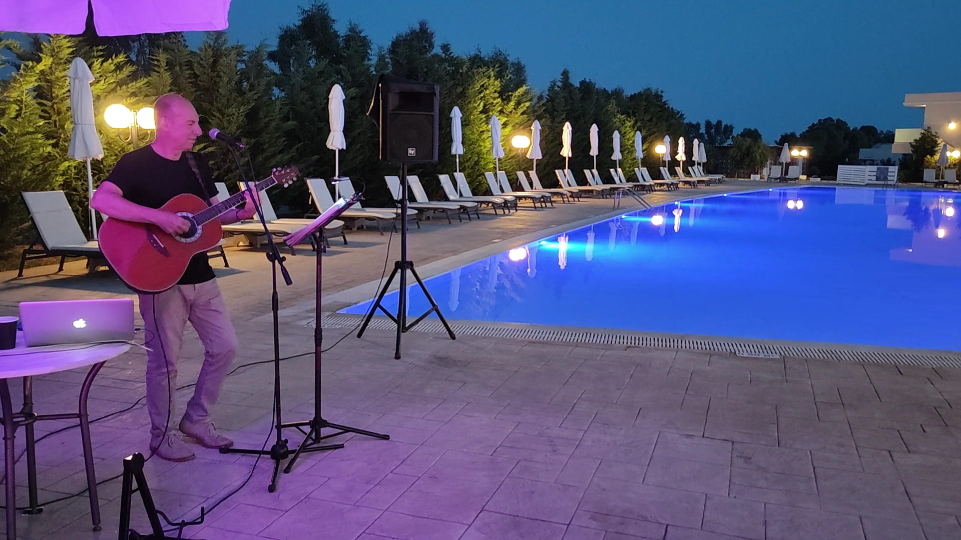 Dzīvās mūzikas vakars Sandy Villas Resort pie izgaismota āra baseina, ģitārists uzstājas vakara stundā pie sauļošanās krēsliem.