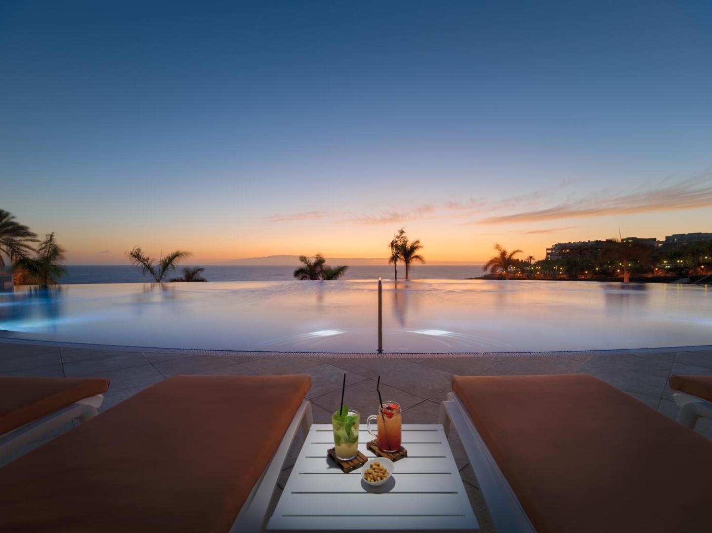 Saulriets pie H10 Atlantic Sunset baseina ar pārplūdes malu Tenerifē — vakara kokteiļi uz galdiņa starp sauļošanās krēsliem