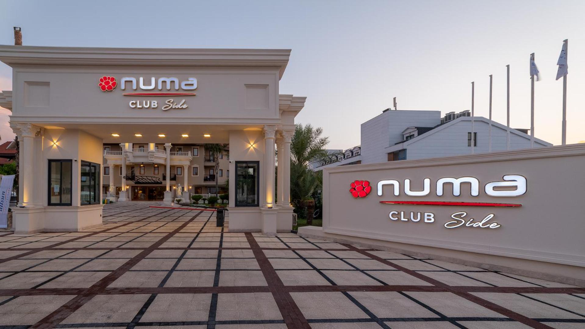 Numa Club Side viesnīcas galvenie vārti un logo pie iebrauktuves dienas noslēgumā ar skatu uz iekšpagalmu