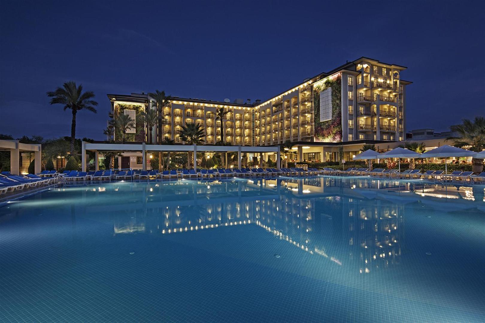 Sunis Elita Beach Resort Hotel & SPA galvenā ēka un izgaismots āra baseins vakarā Sides reģionā, Turcijā