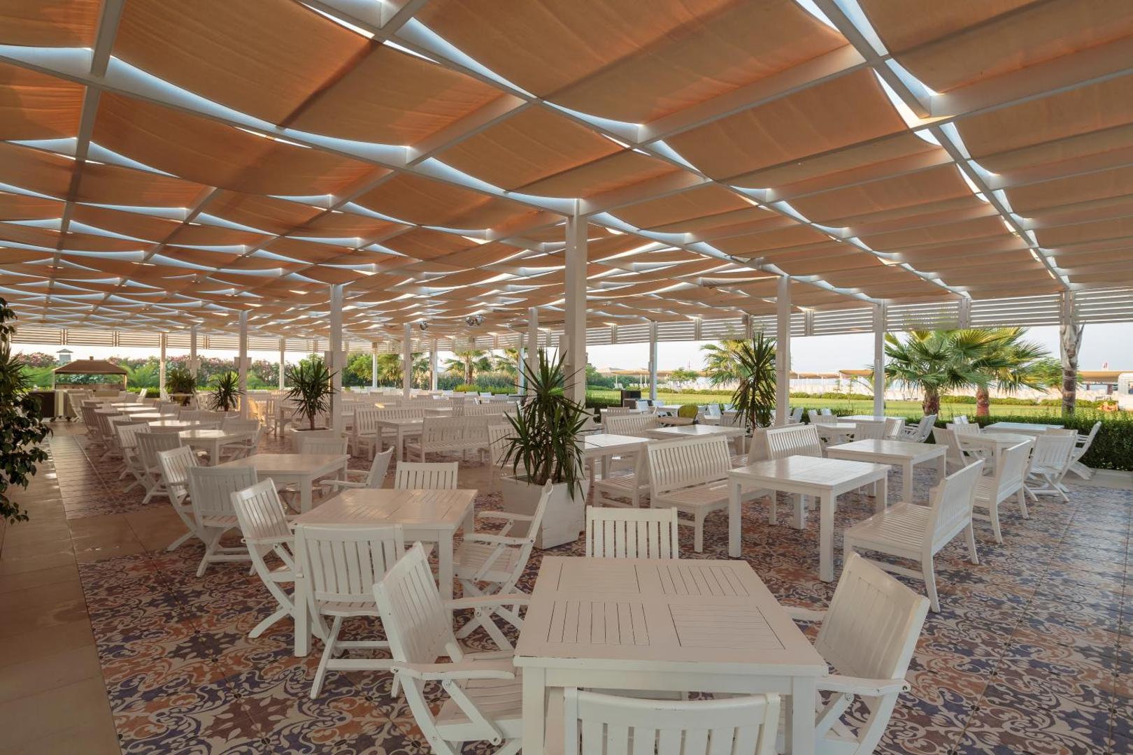 Sunis Elita Beach Resort gaiša āra terase ar baltām mēbelēm un dekoratīvām flīzēm zem saules aizsega pie pludmales