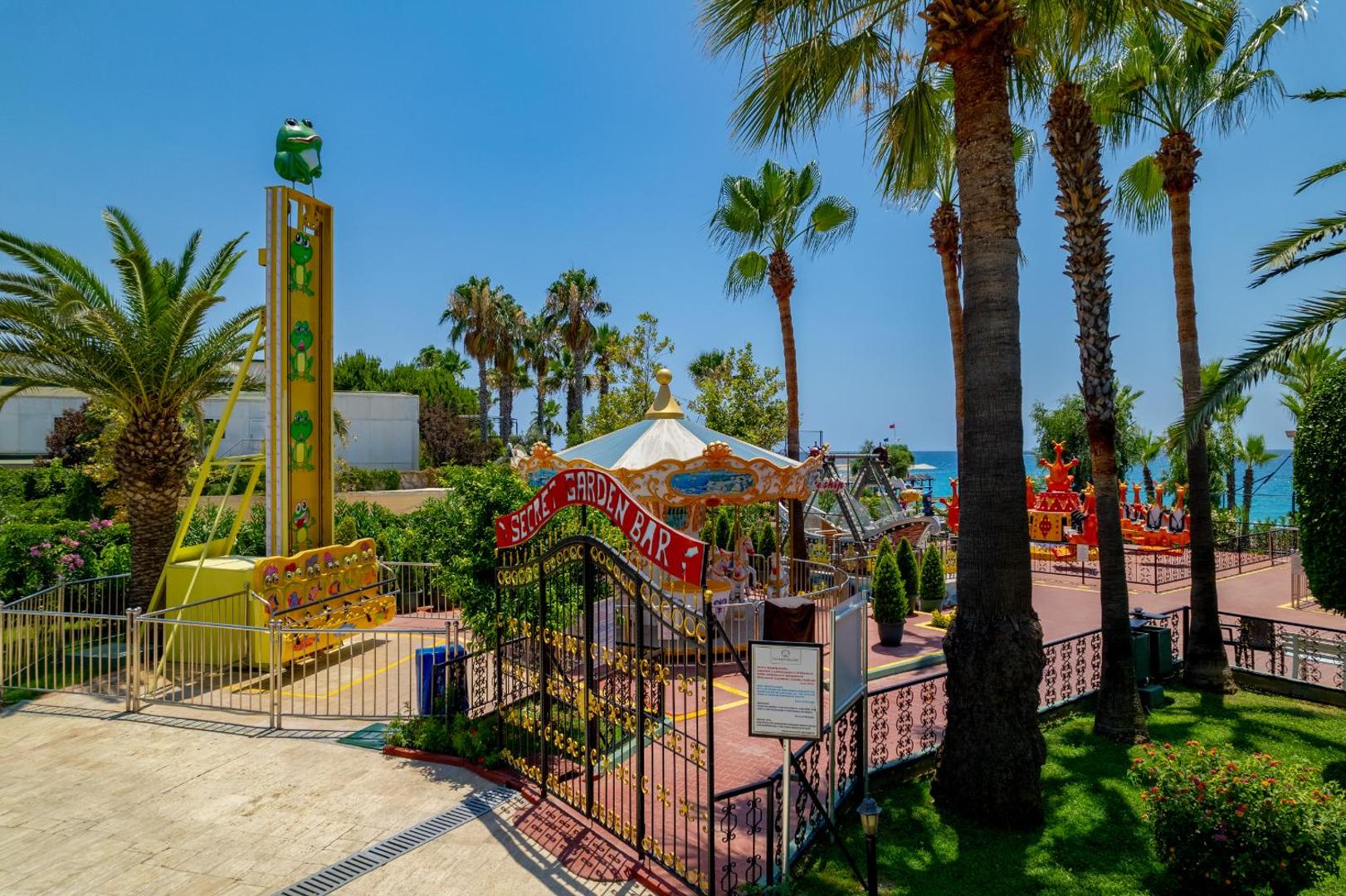 Delphin Deluxe viesnīcas atrakciju parks bērniem ar karuseļiem un palmām Alanjas reģionā, Turcijā — krāsaina izklaides zona pašā jūras piekrastē