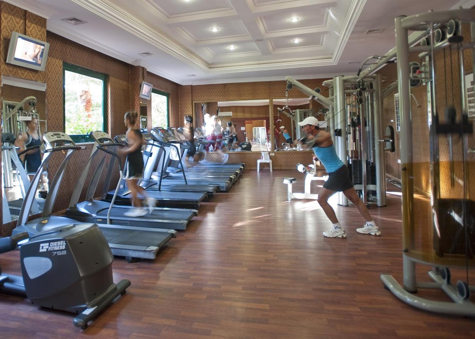 Viesi sporto Delphin Deluxe viesnīcas trenažieru zālē — skrejceliņi un spēka trenažieri plašā telpā ar skatu uz dārzu