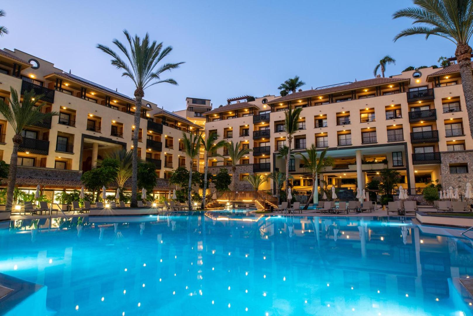 GF Gran Costa Adeje viesnīcas fasāde un baseins vakara apgaismojumā ar izgaismotām palmām un terasēm.