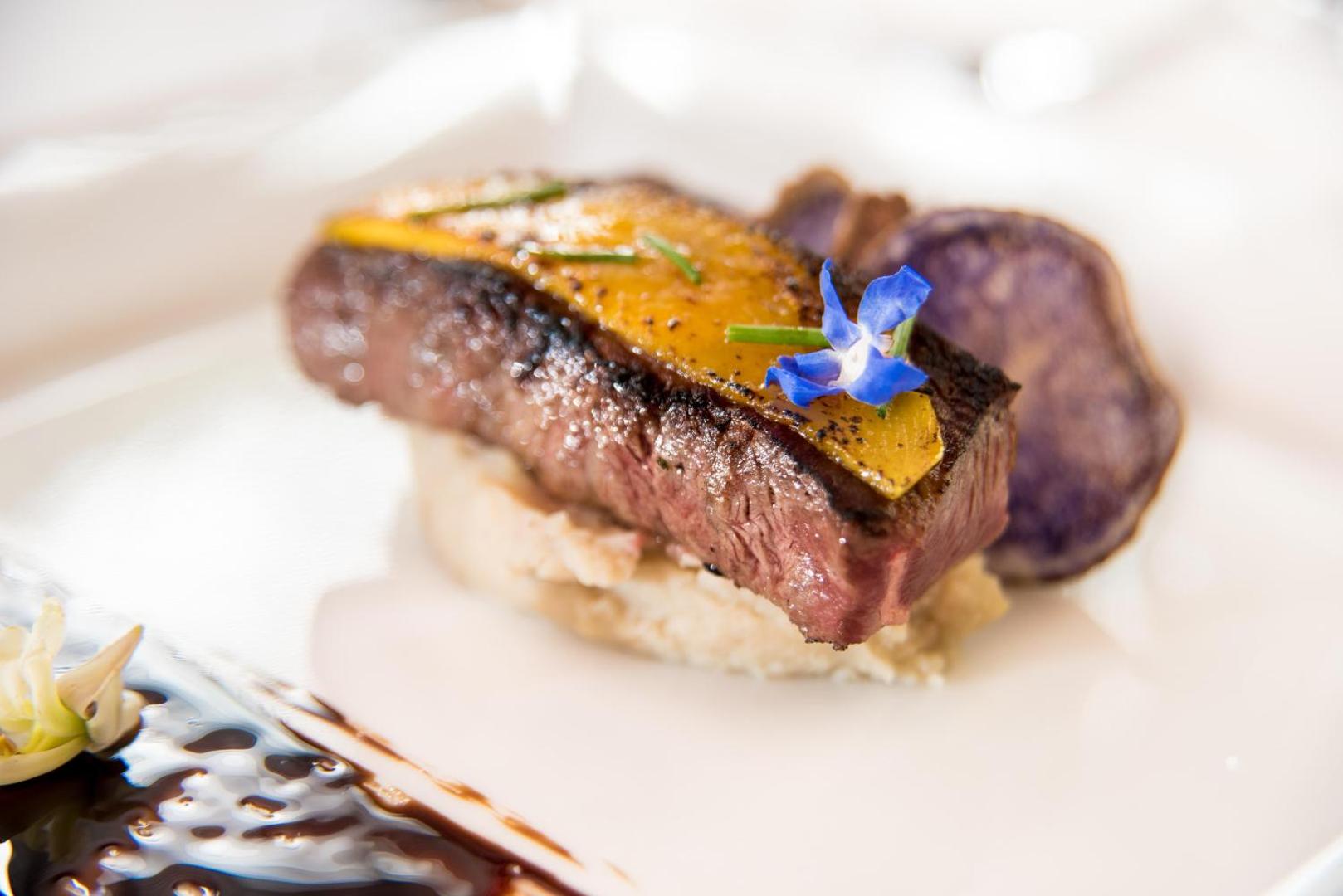 Gourmet restorāna pamatēdiens GF Gran Costa Adeje viesnīcā — eleganti pasniegta gaļa ar dārzeņiem un dekoratīviem ziediem