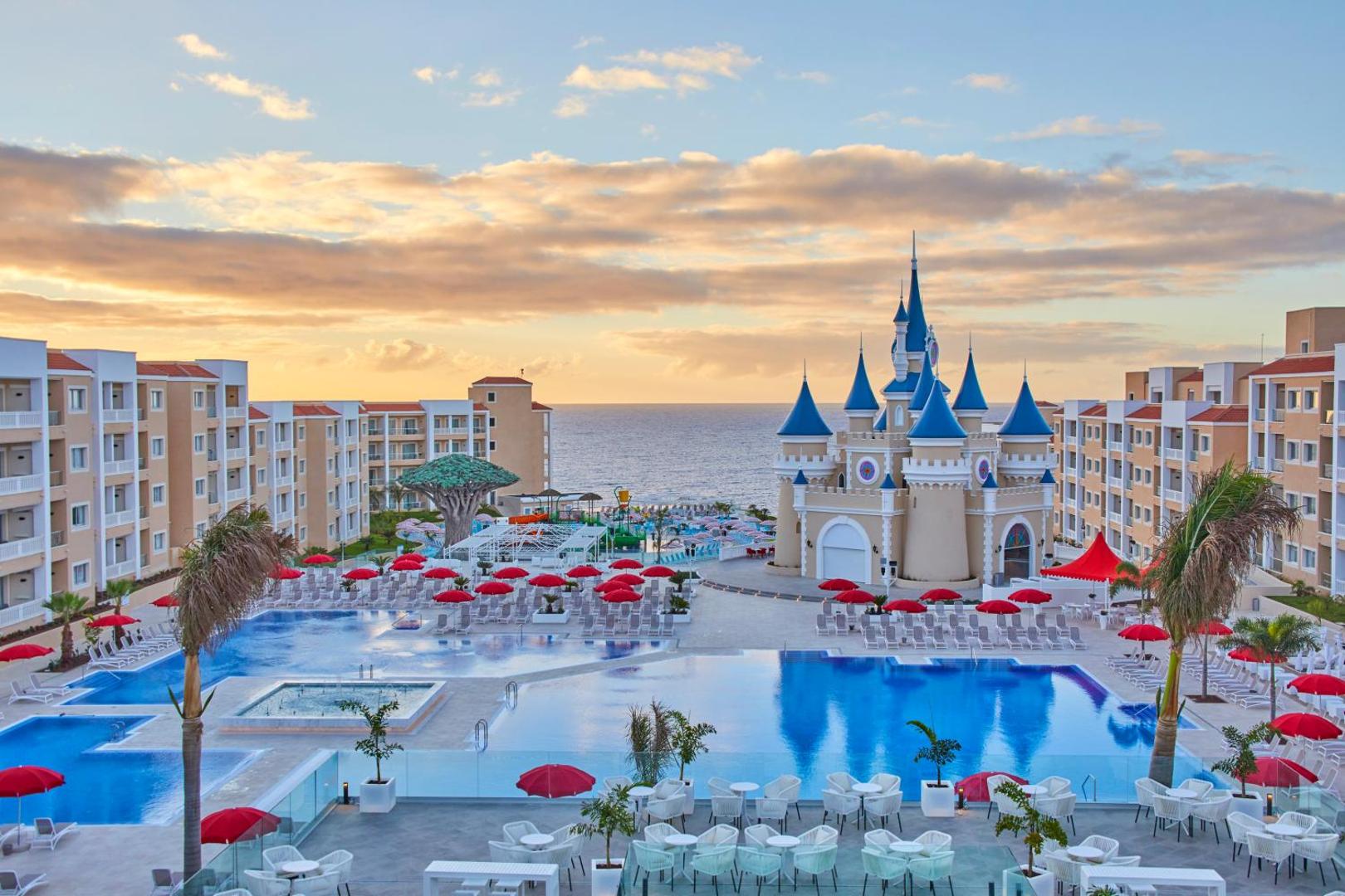 Bahia Principe Fantasia Tenerife kūrorta kopskats saulrietā ar centrālo pili, baseiniem, sarkanajiem saulessargiem un skatu uz jūru Tenerifē.