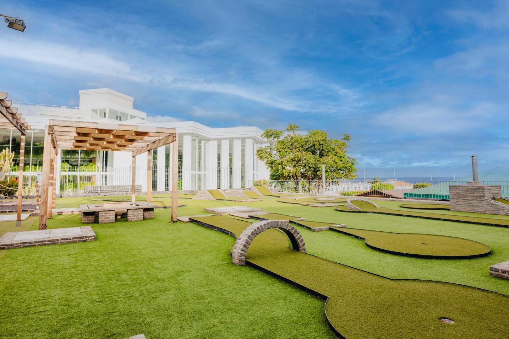 Landmar Playa La Arena minigolfa laukums ar mākslīgo zālienu un šķēršļiem, fonā redzama viesnīcas ēka un okeāna piekrastes ainava.