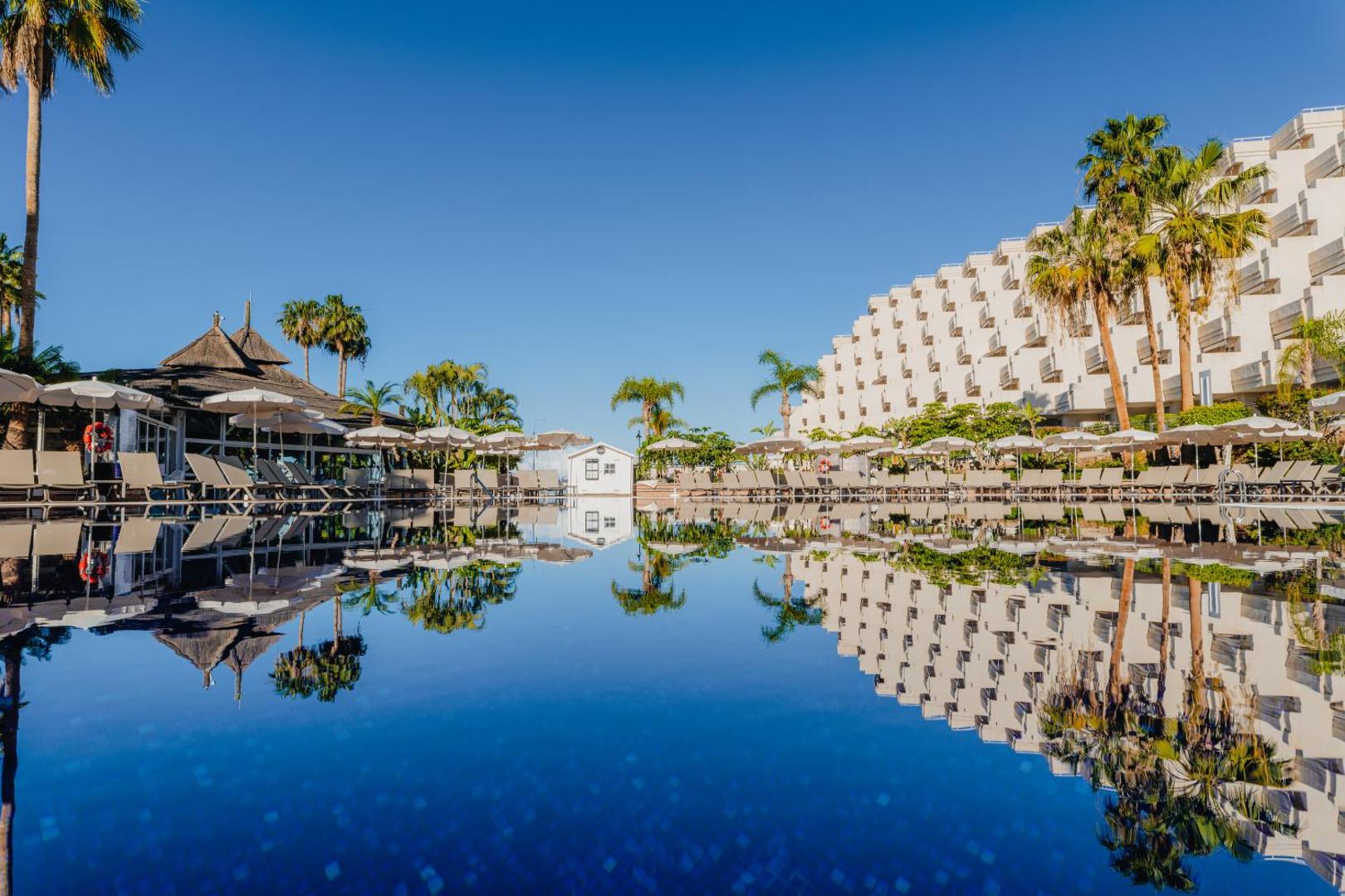 Landmar Playa La Arena viesnīcas galvenais baseins Tenerifē ar ēkas atspulgu ūdenī, saulessargiem un palmām skaidrā dienas laikā
