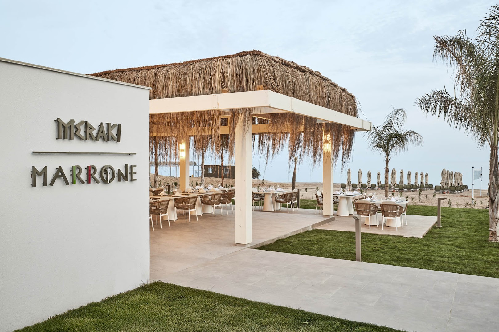 Lusso Sorgun brīvdabas restorāns Meraki Marrone ar salmu jumtu, elegantiem galdiņiem un skatu uz pludmali un palmām.