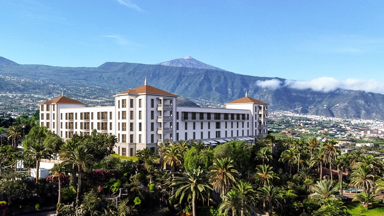 Gran Hotel Taoro ēkas skats no attāluma Puertodelakrusā, Tenerifē — fonā redzams Teides vulkāns un apkārtējie kalni zaļumu ielenkumā
