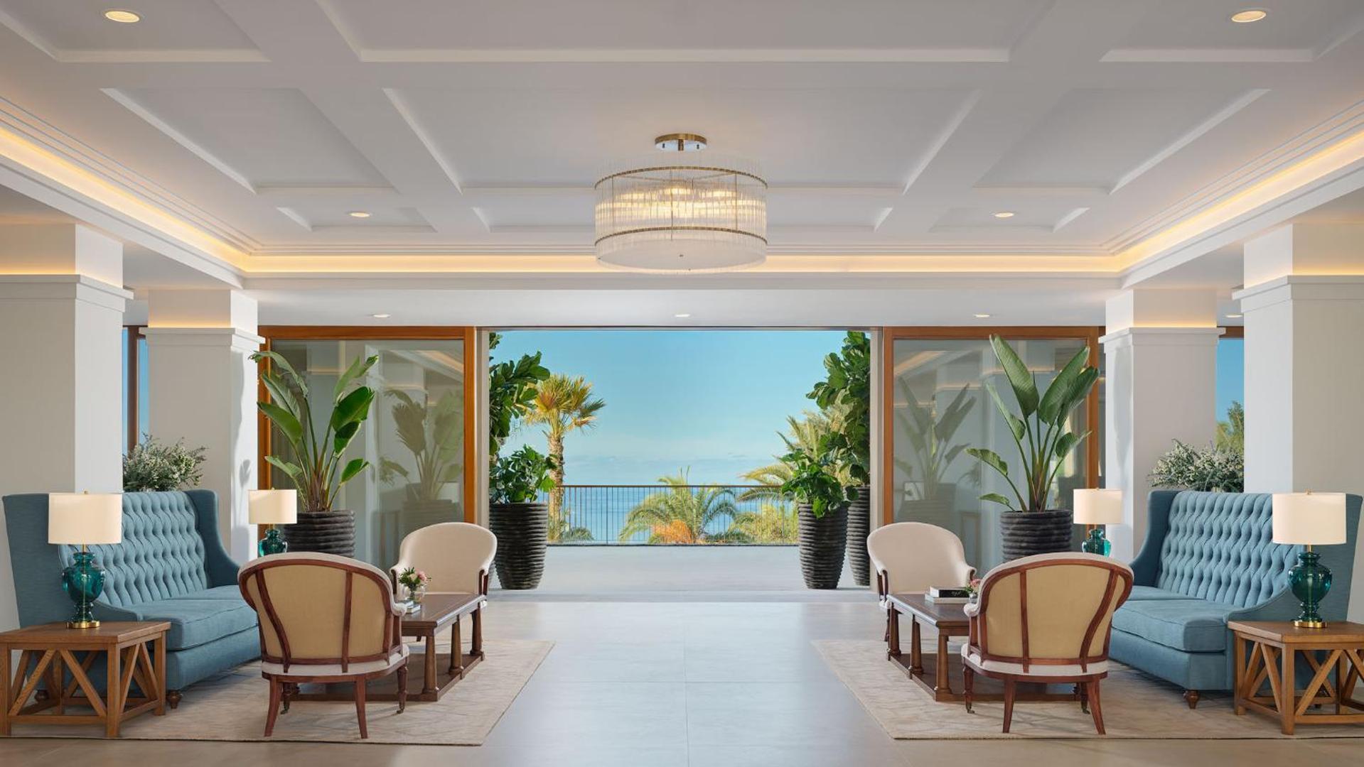 Gran Hotel Taoro greznais vestibils Tenerifē ar gaišām mēbelēm, zilām dīvāniem un skatu uz okeānu caur plašajām terases durvīm