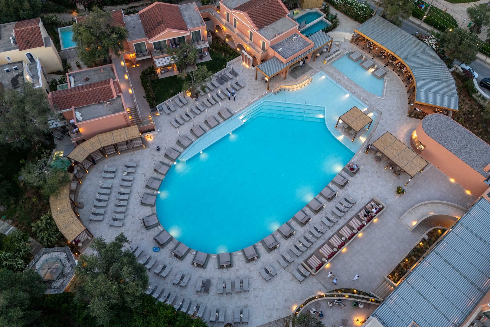 Sentido Apollo Palace viesnīcas teritorija krēslas stundā — izgaismots baseins, bungalo tipa ēkas un olīvkoku dārzs Korfu piekrastē