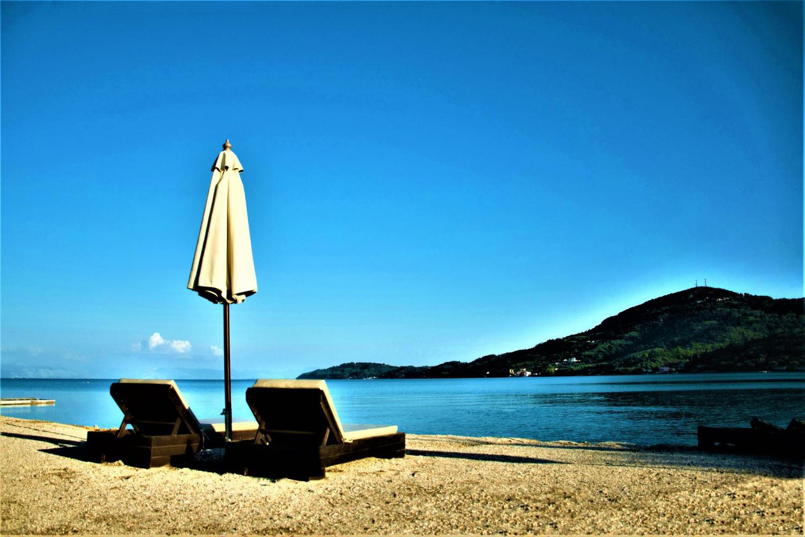 Sentido Apollo Palace privātā pludmale Korfu salā — divi sauļošanās krēsli un saulessargs smiltīs ar skatu uz Jonijas jūru un kalniem