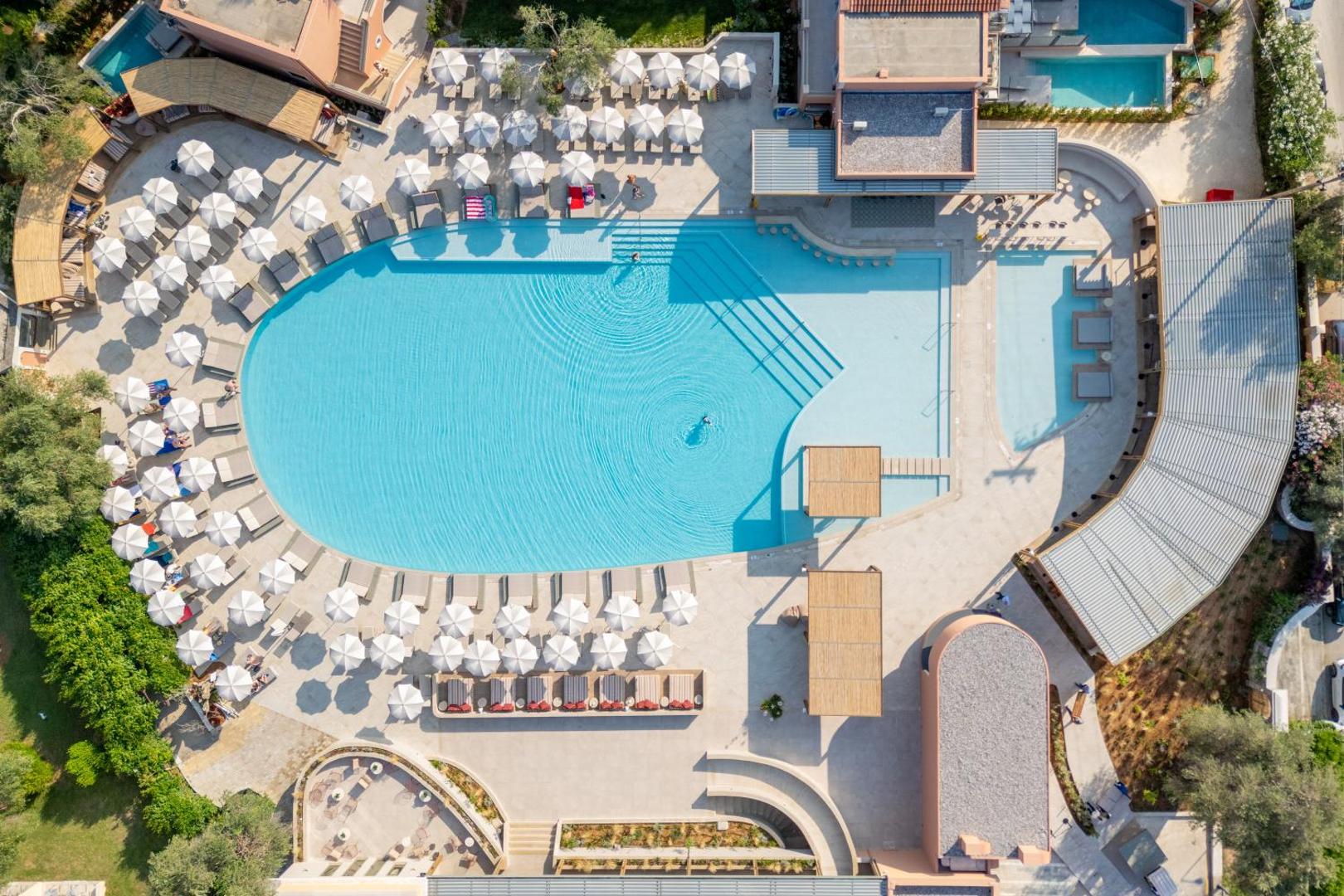 Sentido Apollo Palace viesnīcas galvenais baseins no putna lidojuma Korfu salā — plaša atpūtas zona ar saulessargiem un zviļņiem