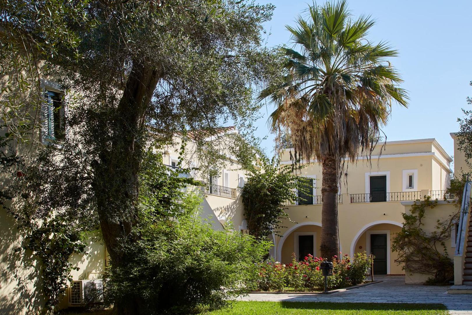 Sentido Apollo Palace viesnīcas iekšpagalms ar palmu un ziedošām rozēm — tradicionāla grieķu arhitektūra ar arkām un balkoniem