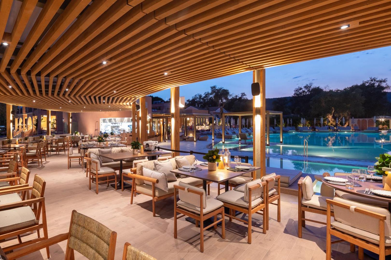 Sentido Apollo Palace modernais restorāns vakarā ar koka siju griestiem, mīkstajiem krēsliem un skatu uz izgaismoto āra baseinu