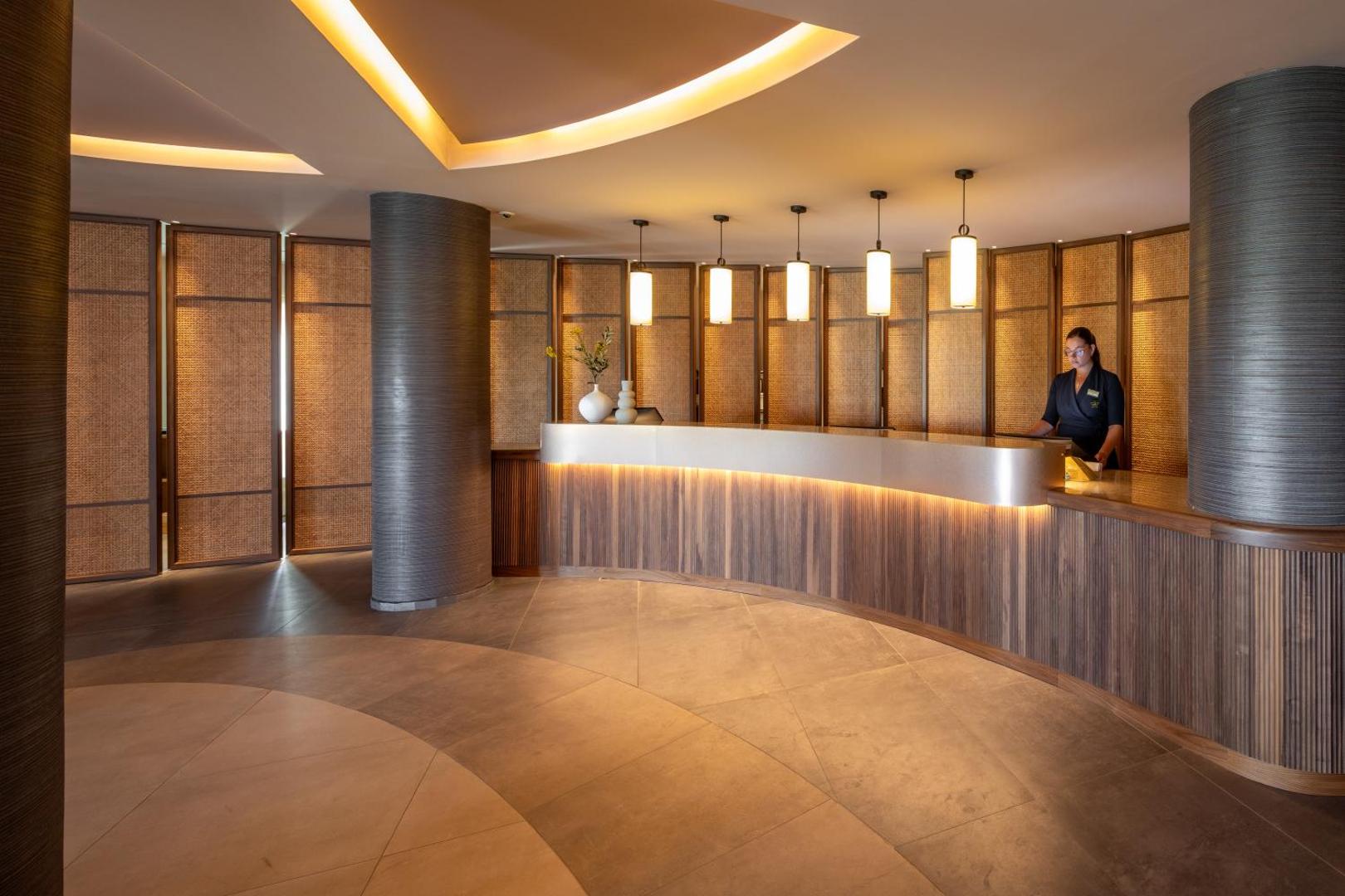 Sentido Apollo Palace viesnīcas recepcijas lete ar koka apdari un siltu apgaismojumu — viesu uzņemšanas zona Korfu salā