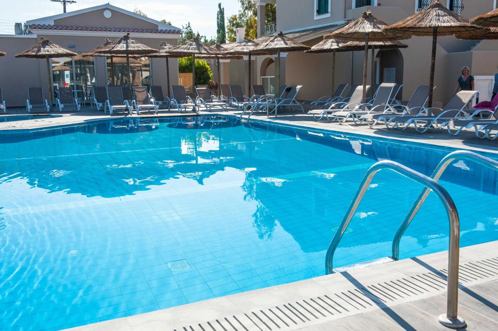 Amour Holiday Resort galvenā baseina tuvplāns ar kāpnēm, sauļošanās krēsliem un salmu saulessargiem saulainā dienā Korfu p...