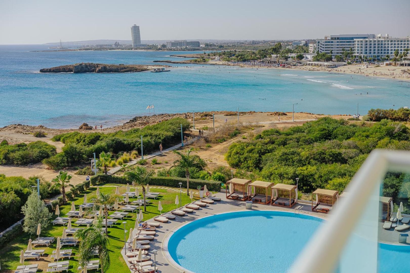 NissiBlu Beach Resort skats no augšas uz āra baseinu, sauļošanās zonu un Nissi pludmali Ajanapā, Kiprā — palmas un jūra