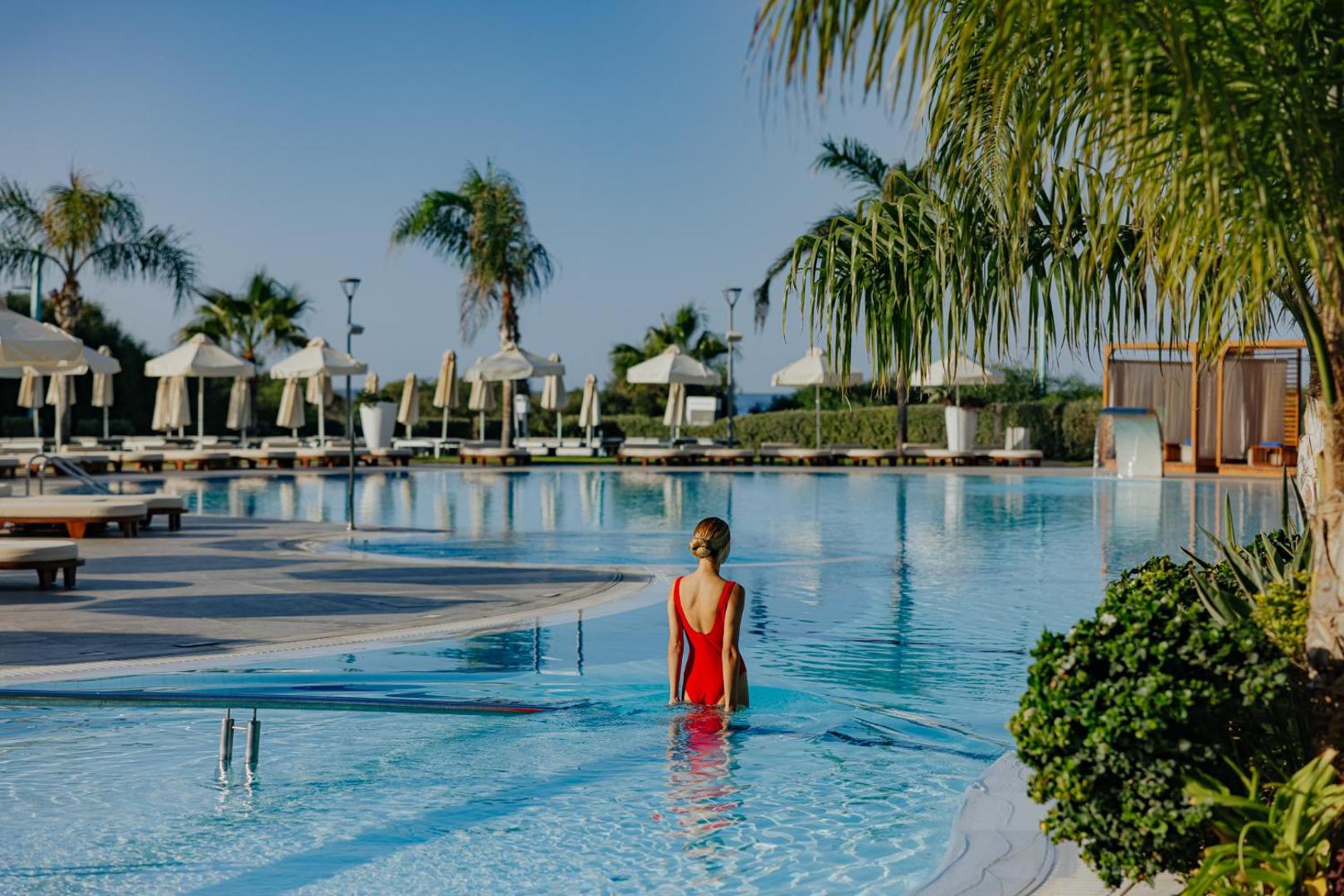 NissiBlu Beach Resort āra baseins ar saulessargiem un atpūtas krēsliem — sieviete sarkanā peldkostīmā ieiet dzidrā ūdenī