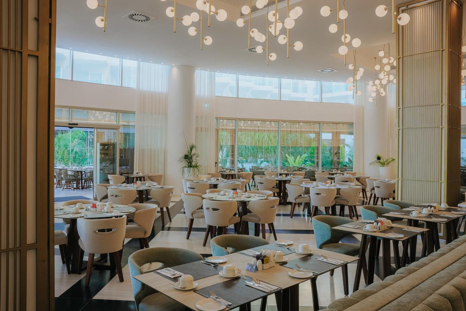 Nissiblu Beach Resort galvenais restorāns ar modernu interjeru, gaišām mēbelēm un dizaina lustrām — plaša un saulaina brokastu zona