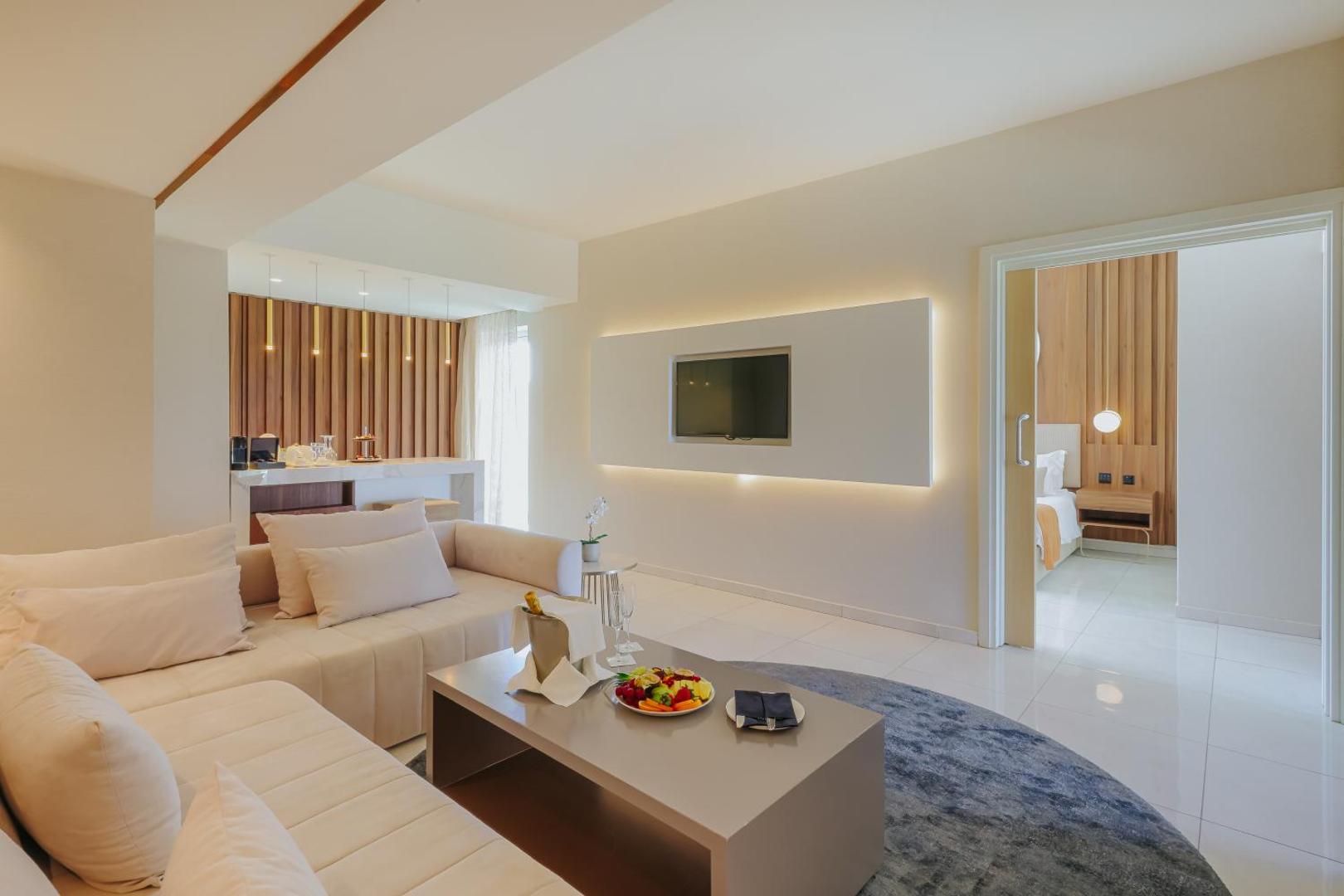 NissiBlu Beach Resort numura viesistabas zona ar mīksto stūra dīvānu, TV un augļu plati — elegants interjers gaišos toņos