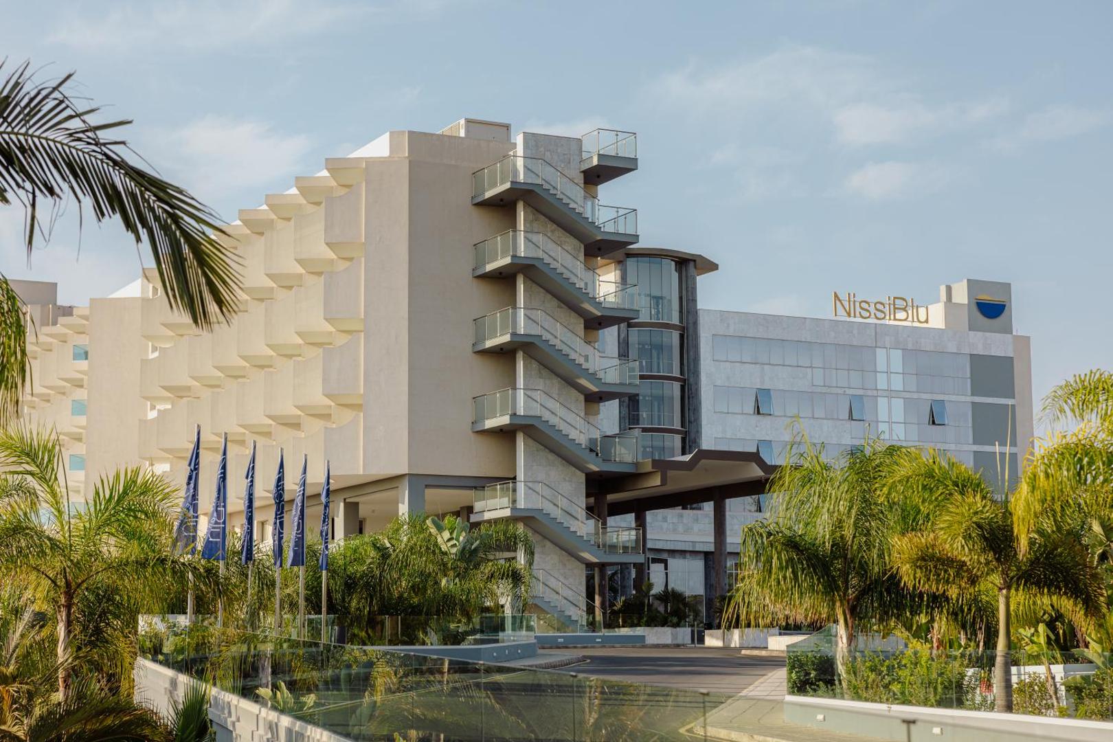 Nissiblu Beach Resort modernā arhitektūra un galvenā ieeja Aijanapā — stiklota fasāde, palmas un karogi pie pieczvaigžņu viesnīcas