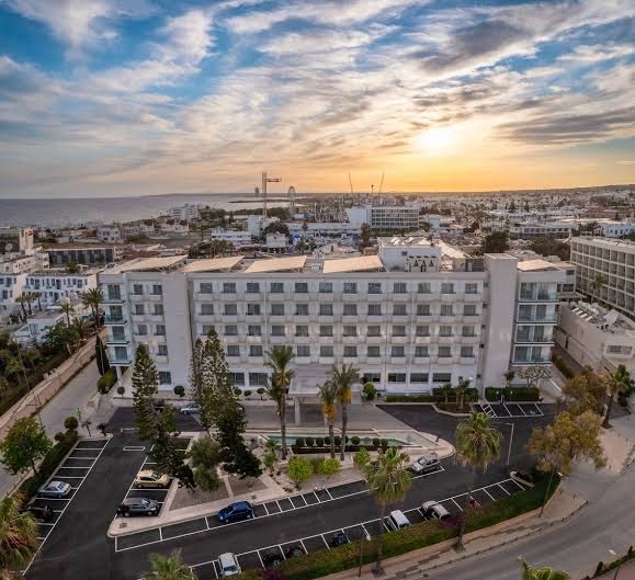Nestor Hotel skats no putna lidojuma saulrietā Aijanapā, Kiprā — viesnīcas fasāde, autostāvvieta un pilsētas panorāma pie jūras