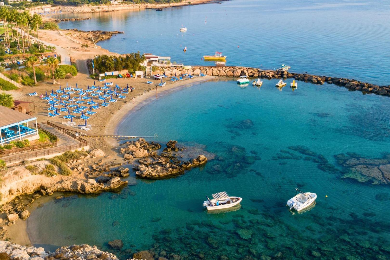 Cavo Maris Beach pludmales līcis ar kristāldzidru tirkīza ūdeni Protarasā — redzamas laivas, klintis un zili saulessargi smiltīs