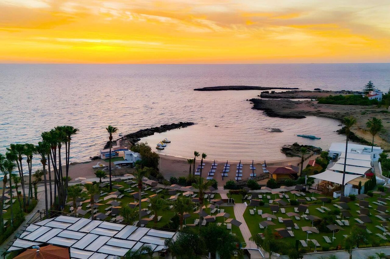 Cavo Maris Beach viesnīcas skats no augšas uz privāto līci saulrietā Protarasā, Kiprā — pludmale, palmas un saulessargi zeltītā gaismā