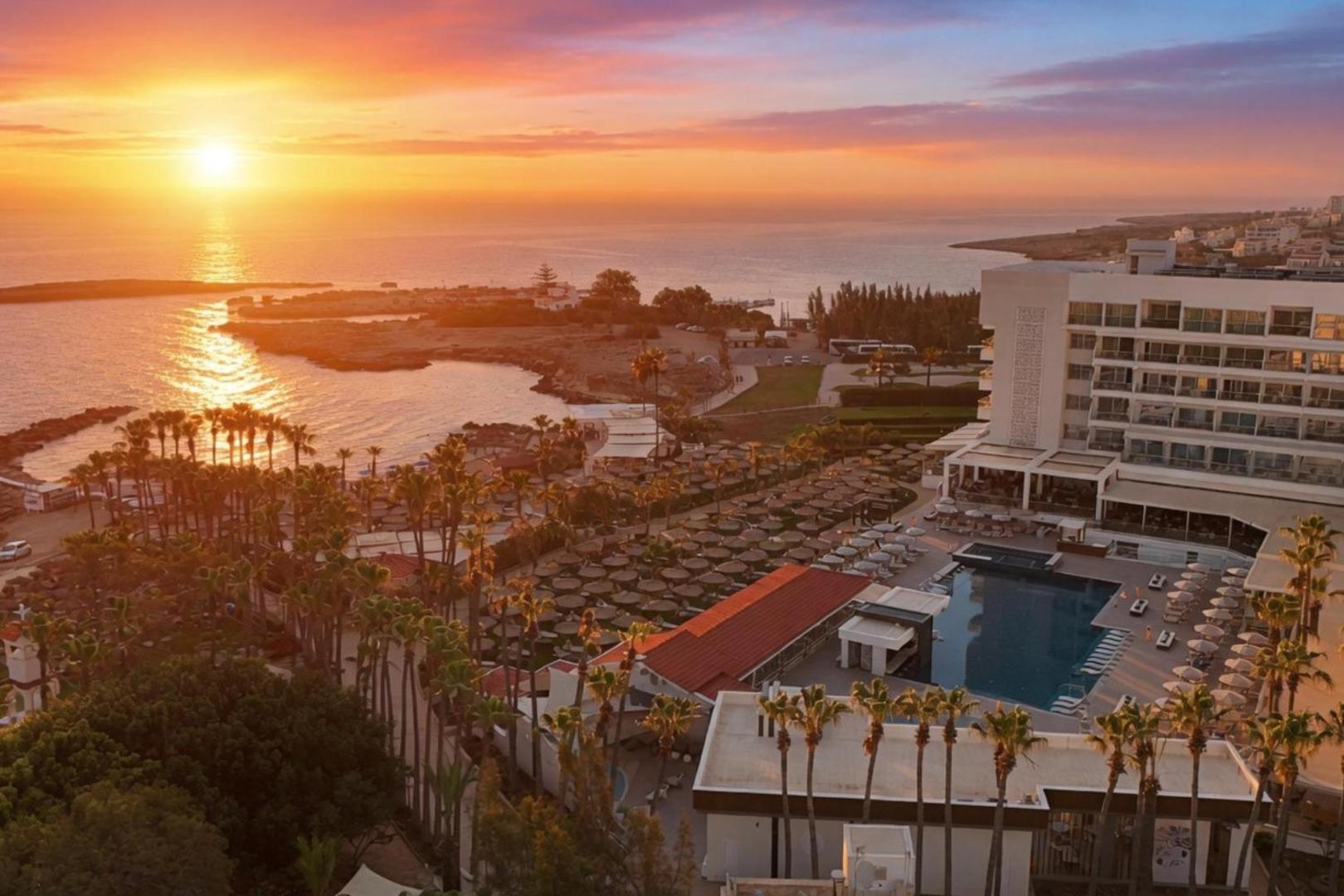 Cavo Maris Beach Hotel skats no augšas saulrietā Protarasā, Kiprā — baseins, palmas un Vidusjūras piekraste vakara gaismā