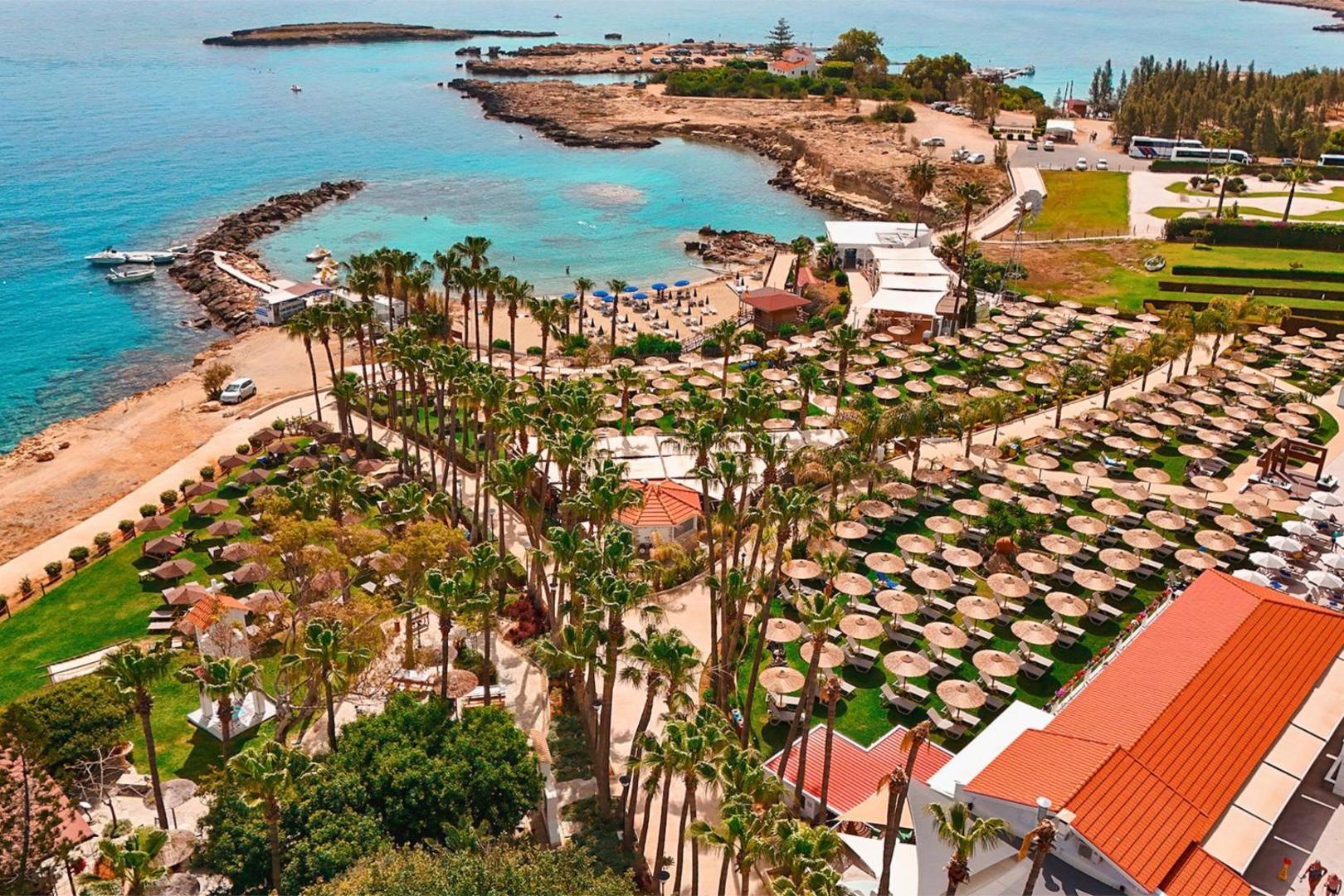 Cavo Maris Beach viesnīcas teritorijas panorāma no augšas — plašs dārzs ar simtiem saulessargu, palmas un pieeja tirkīza līcim